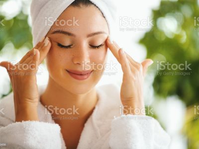 Young woman applying and rubbing anti age cream with her eyes closed. She's in the bathrobe and her hair is wrapped in the towel.