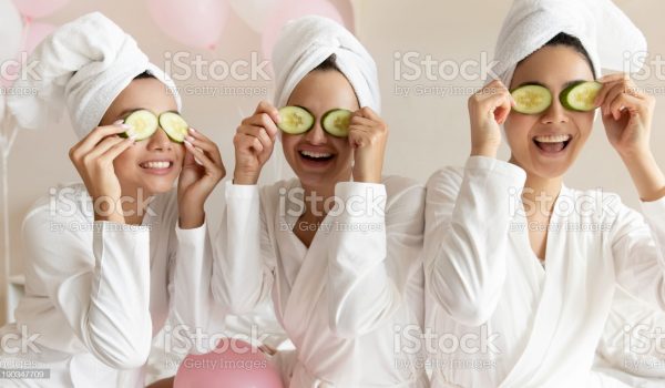 Happy young women wear white bathrobes towels on head make cucumber facial skin care mask on eyes laughing relaxing together, smiling girls friends having fun on spa beauty salon party with balloons