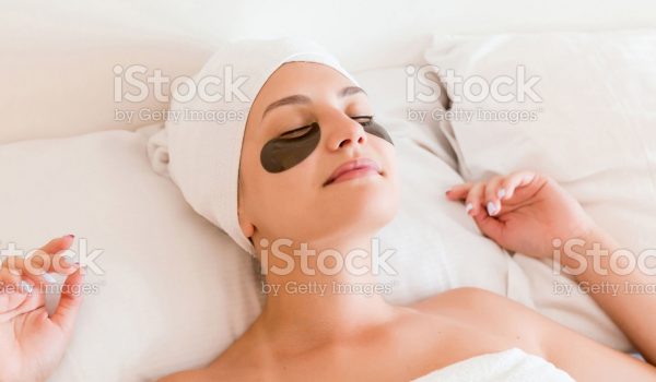 Young woman with closed eyes and black eye patches is lying and relaxing in the bed after having a bath wrapped in towel. Beauty treatment and skincare concept.