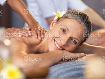 Happy senior woman getting oil massage on her back at spa center. Portrait of senior woman receiving back massage. Closeup face of smiling mature woman relaxing on massage table and looking at camera.